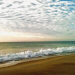 Ocean waves rolling onto a sandy beach beneath a textured cloud-filled coastal sky